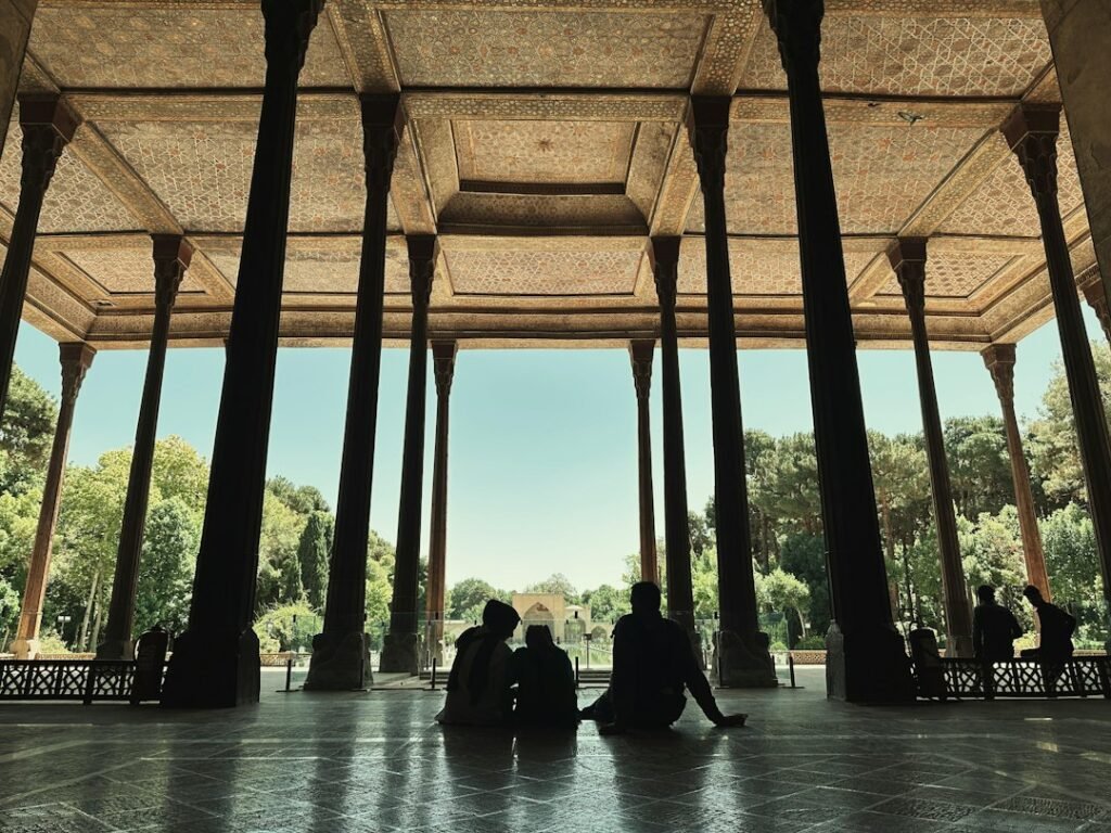 Visiteurs assis à contre-jour sous les hautes colonnes d’un pavillon persan ouvert sur un jardin
